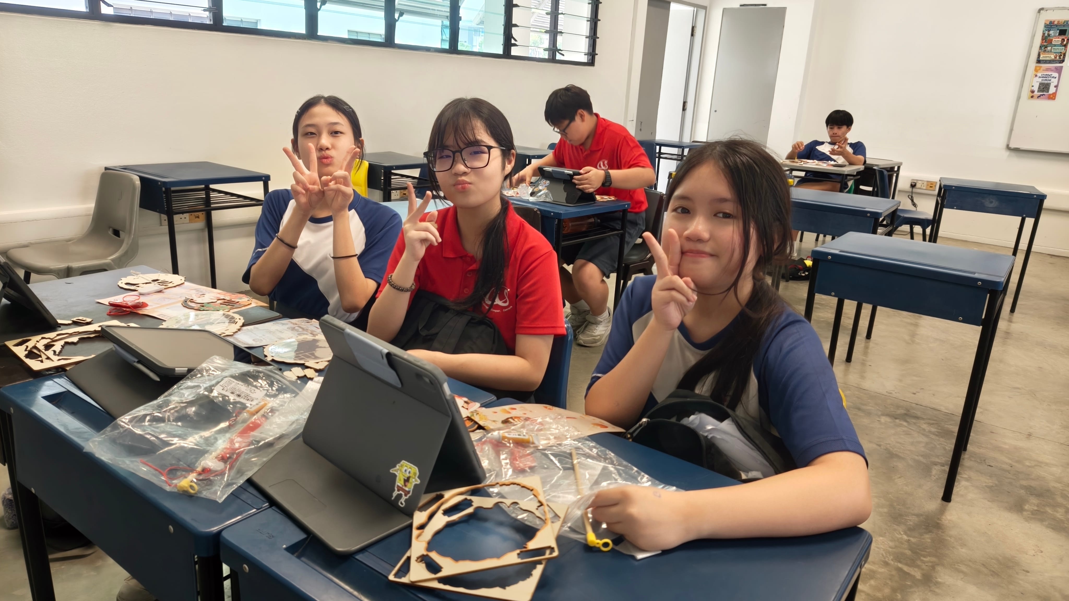 Three secondary school girls sit at their desks in a classroom, smiling and giving peace signs at the camera. Their desks are covered with wooden Mid-Autumn Festival lantern craft kit pieces and tablets. Two boys can be seen working at their desks in the background.